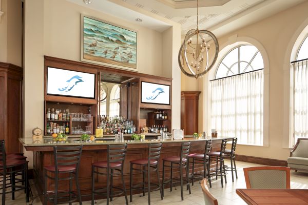 A stylish bar with high chairs, bottles, and decor. There are screens above and large windows in the background.