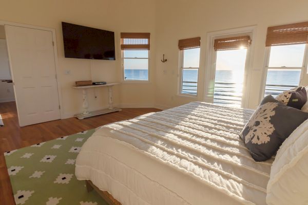 A bright bedroom with ocean views through windows, a bed with white linens, a wall-mounted TV, and a decorative table underneath.