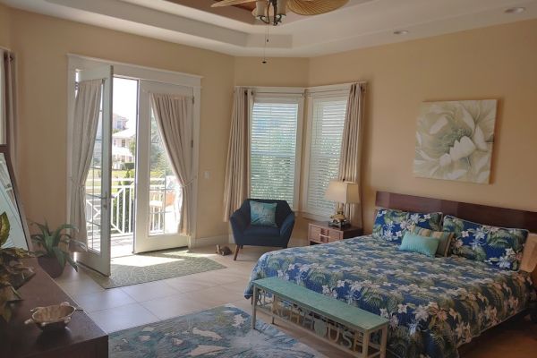 A cozy bedroom with a floral bedspread, large window, open doors leading to a balcony, armchair, and tropical decor for a relaxed vibe.