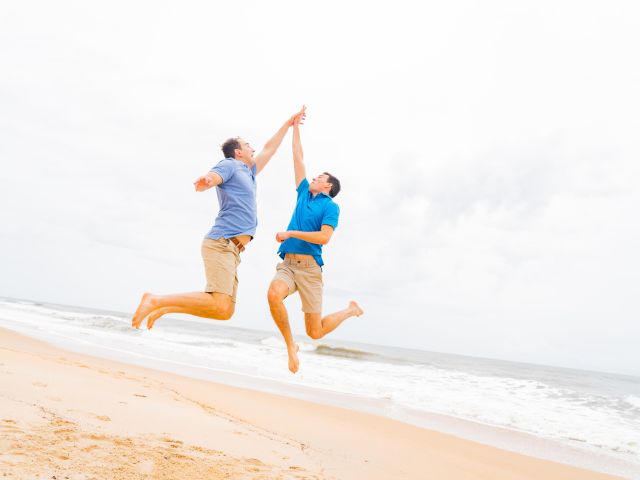 Two people in blue shirts and shorts are jumping and high-fiving on a sandy beach with waves in the background.